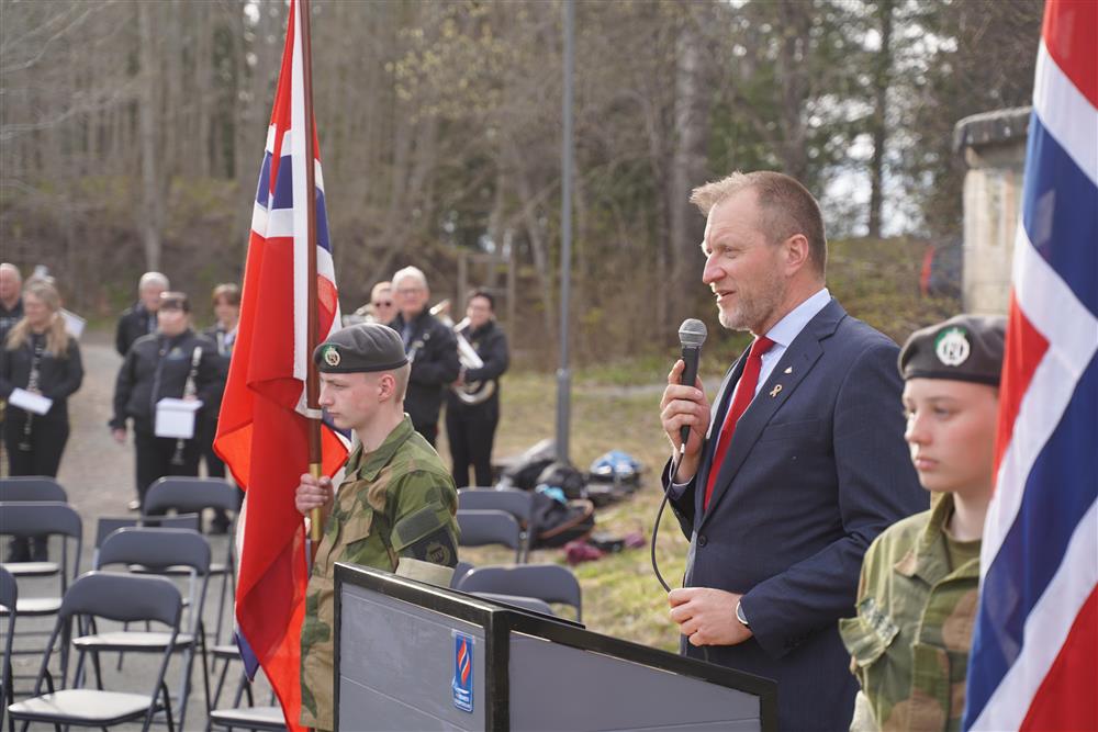 En mann står bak et podium og taler, på hver side står det en flagbærer, i bakgrunnen står korpset. - Klikk for stort bilde