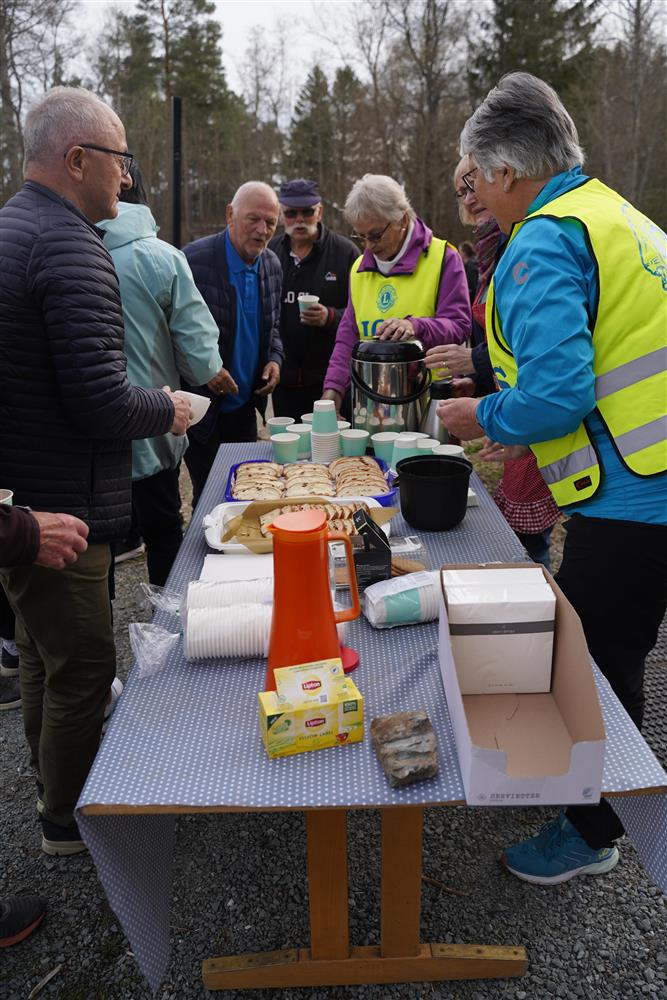 Flere personer rundt et bord utendørs. Bordet er fult av kaker og kaffe. Tre damer med refleksvest gir ut kake og kaffe. - Klikk for stort bilde