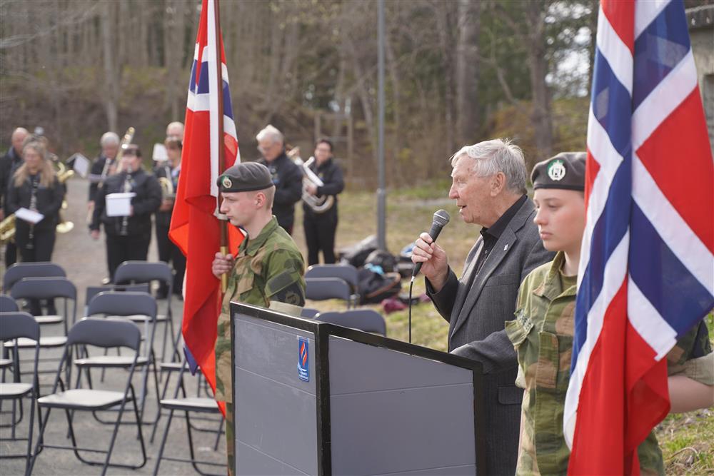 En mann står bak podiumet og holder en tale, på hver side står en flaggbærer, i bakgrunnen står korpset. - Klikk for stort bilde