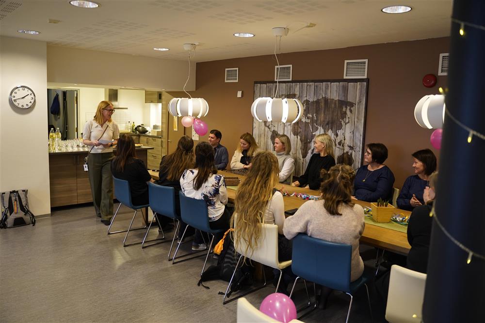 Mange mennesker som sitter rundt bordet inne på Ungdomskontakten, Sissel står ved enden og taler. - Klikk for stort bilde