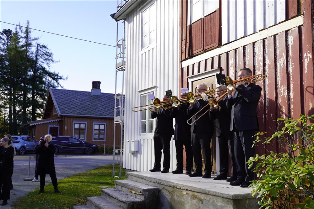Flere mennesker som spiller ulike blåseinstrumenter i sola, de står oppå en trapp like ved senteret. - Klikk for stort bilde