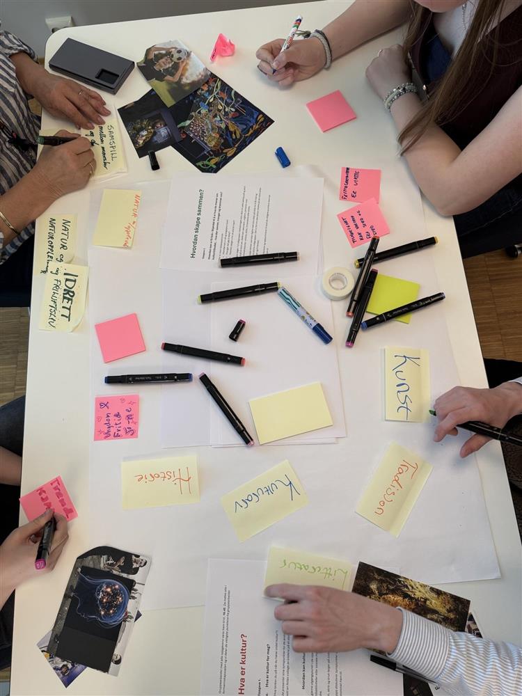 Mange forskjellige lapper og notater som er spredd utover et bord, fire personer sitter rundt bordet og skriver. - Klikk for stort bilde