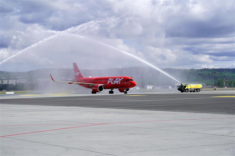 Et rødt fly med bokstavene &quot;Play&quot; på siden, det er sett på avstand og kjører gjennom en bue av vann. Det er sol og litt skyer. - Klikk for stort bilde