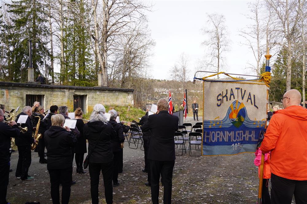 Skatval hornmusikklag spiller. De står foran podiumet, til høyre står en mann med korpset sin vimpel. - Klikk for stort bilde