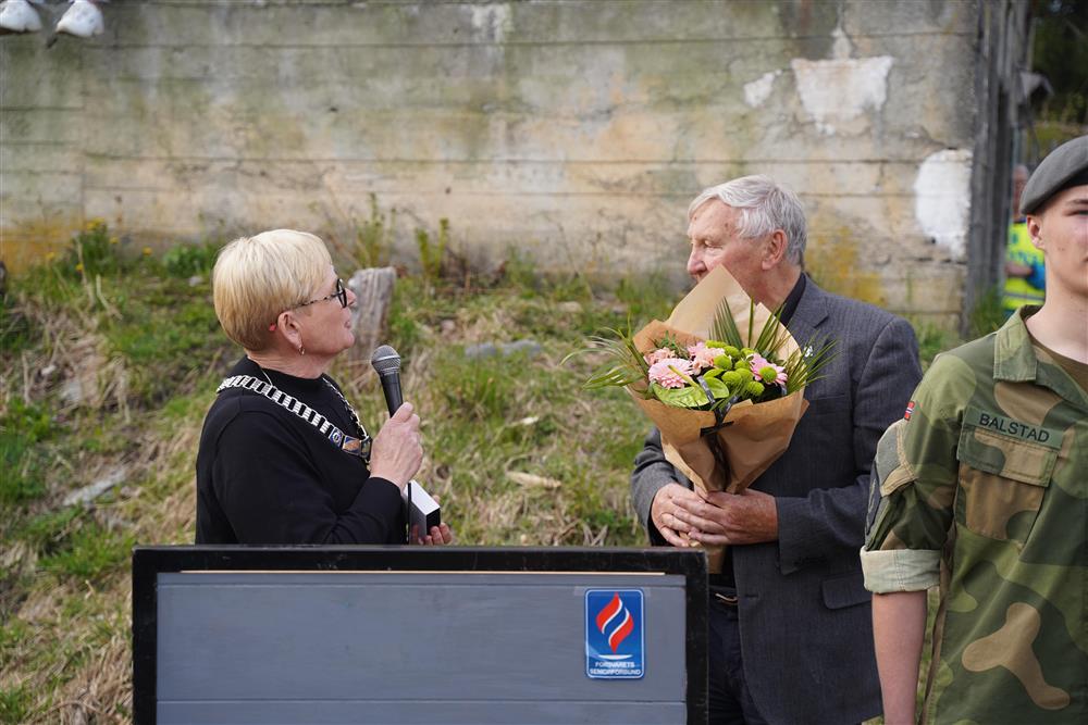 Ordførern gir blomster til Ole Myrbekk. De står begge bak podiumet og snakker til hverandre. Ole holder blomstene, ordføreren har en mikrofon. - Klikk for stort bilde