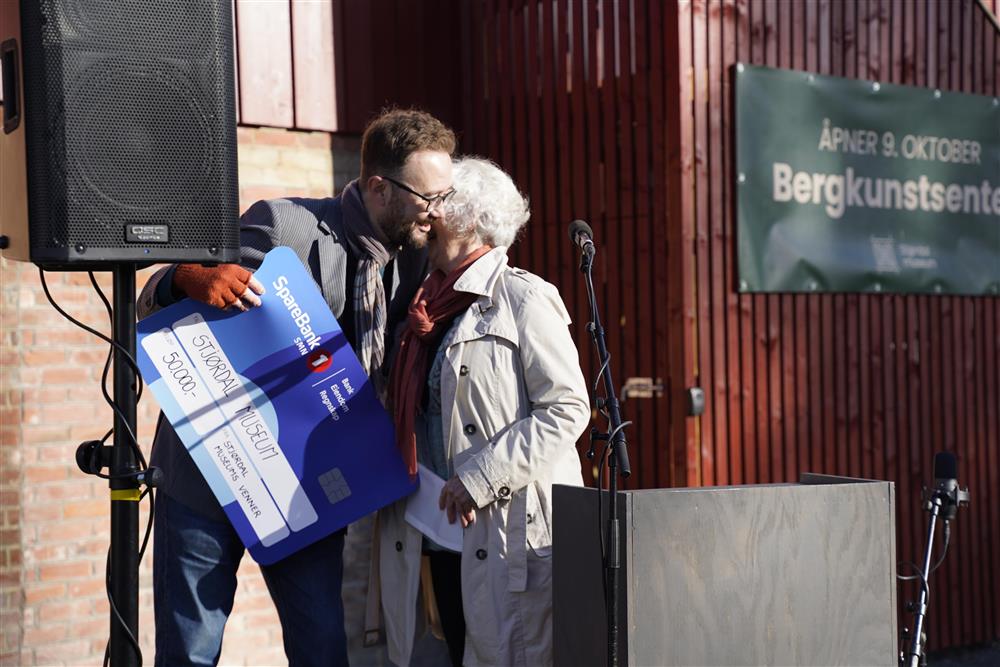 En mann som tar imot en stor sjekk fra en eldre dame, de klemmer. - Klikk for stort bilde