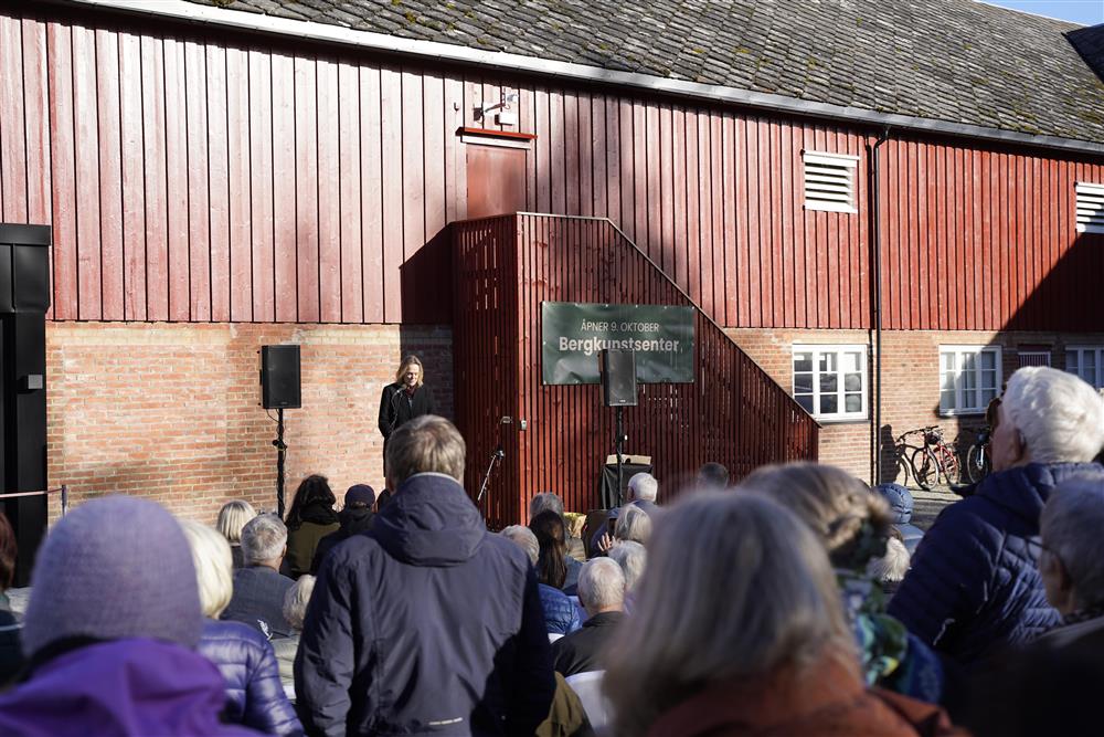 En dame som står foran et stort publikum å taler, hun står på en scene rigget opp utenfor senteret. Noen folk sitter, mens andre står. - Klikk for stort bilde