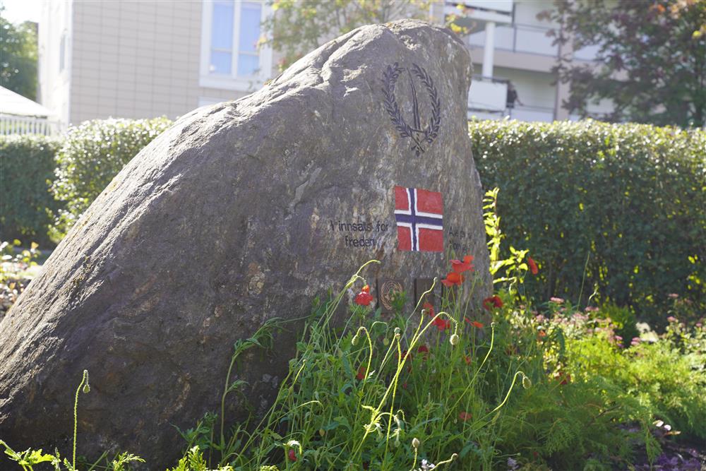 Minnesteinen med norsk flagg og andre viktige symboler, foran er det plantet blomster. - Klikk for stort bilde