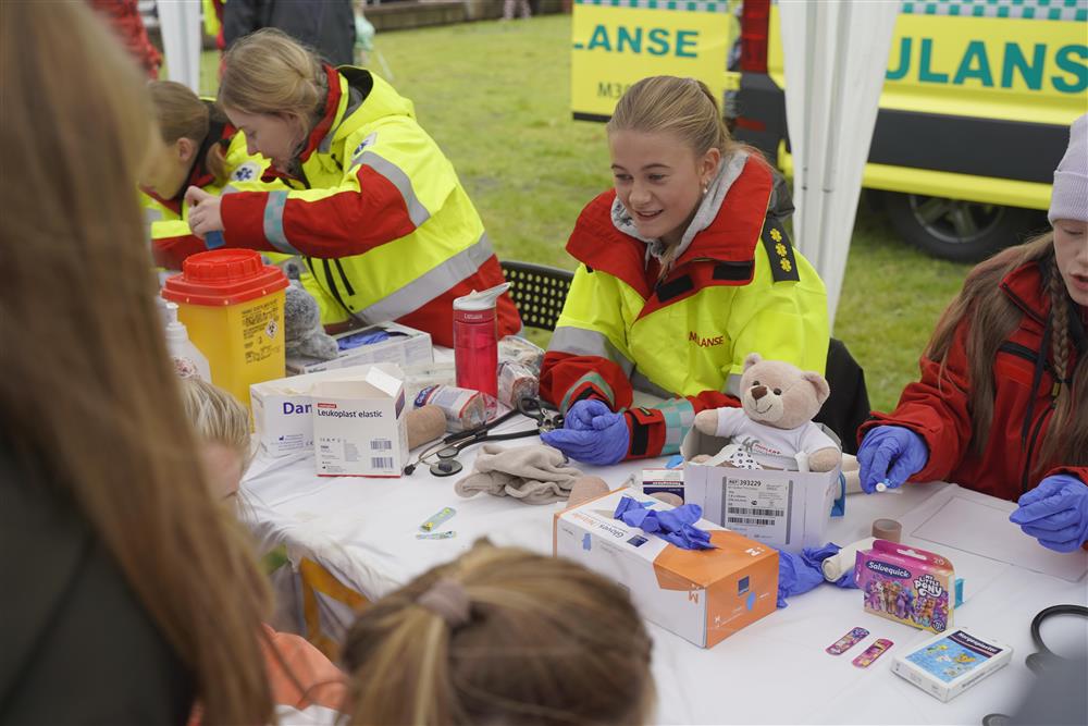 Flere barn som har levert inn bamsene sine til ambulanse arbeidere som sitter bak bord, de tar på plaster, lytter med stetoskop og mer. - Klikk for stort bilde