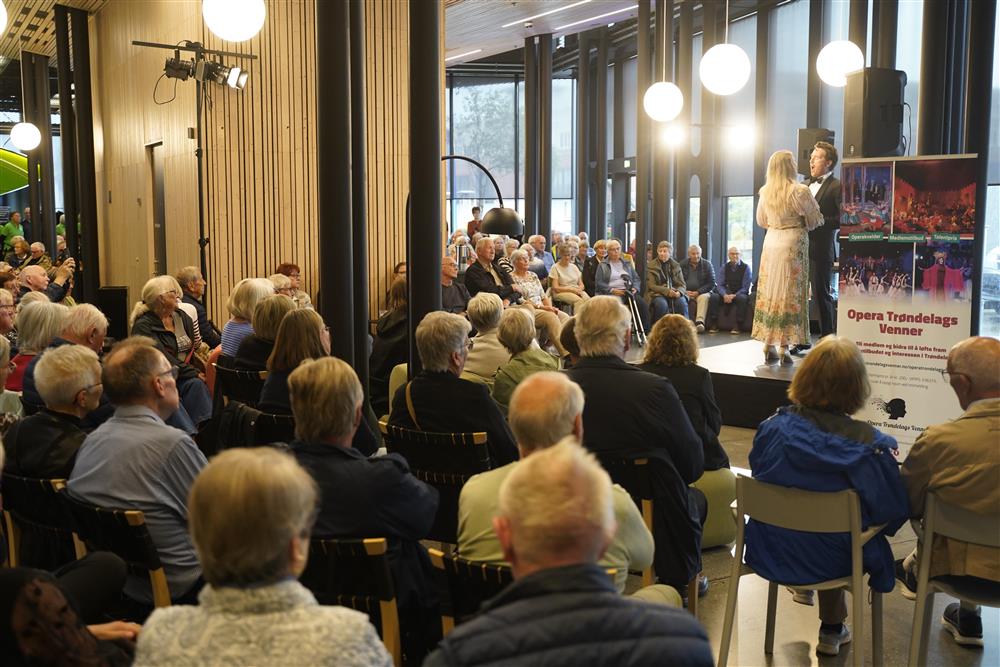 En mann og en dame på scenen i foajeen, det har blitt enda mer publikum og har måttet settes fram ekstra stoler. Noen står og noen sitter også på gulvet. - Klikk for stort bilde