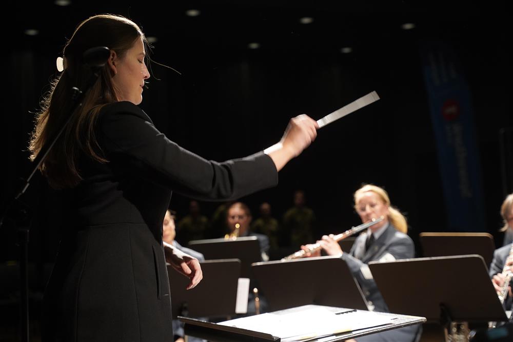 En kvinnlig dirigent som dirigerer, i bakgrunnen spiller flere folk instrumenter. - Klikk for stort bilde