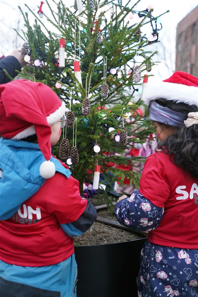 To barnehage barn med nisseluer som henger på julepynt - Klikk for stort bilde