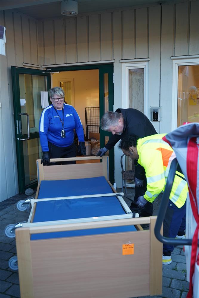 Flere personer som transporterer en seng. De er utendørs. - Klikk for stort bilde