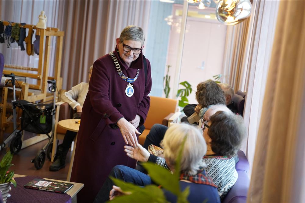 Ordføreren som håndhilser på en person som sitter i en sofa, det sitter flere personer i sofa og stoler rundt - Klikk for stort bilde