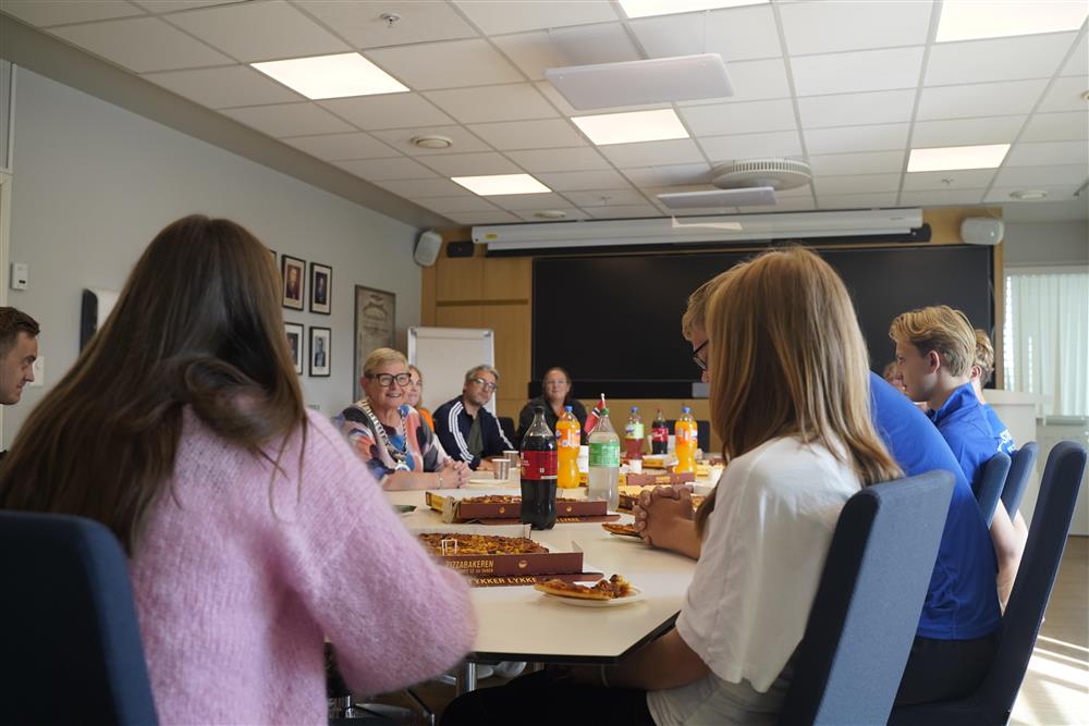 Ungdomsutøvere, ordføreren samt frivillige sitter rundt bord i formannskapssalen, de spiser pizza og drikker brus. - Klikk for stort bilde