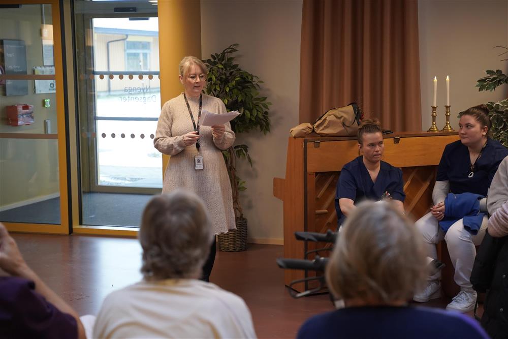 En dame som leser fra en lapp og flere folk som ser på - Klikk for stort bilde