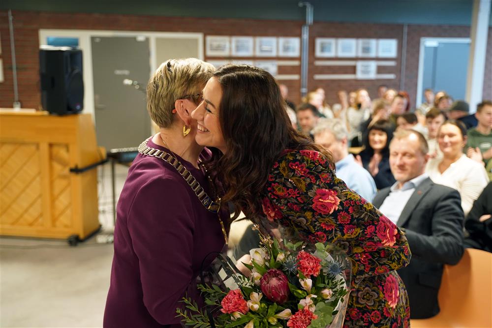 Ordfører Eli Arnstad overrekker blomster til rektor Rachel Johansen. De klemmer hverandre og smiler, det skjer innendørs i kantina foran et publikum - Klikk for stort bilde