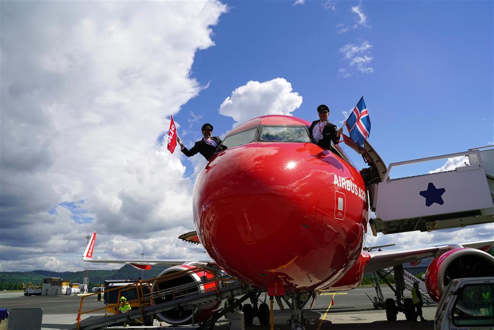 To piloter som stikker hode ut av hvert sitt vindu på et Airbus fly. Den til venstre holder et flag med logoen til flyselskapet Play, den andre et Islandsk. Det er blå himmel med litt sky bak dem, under flyet holder folk på å gjøre klar til at flyet skal dra.  - Klikk for stort bilde