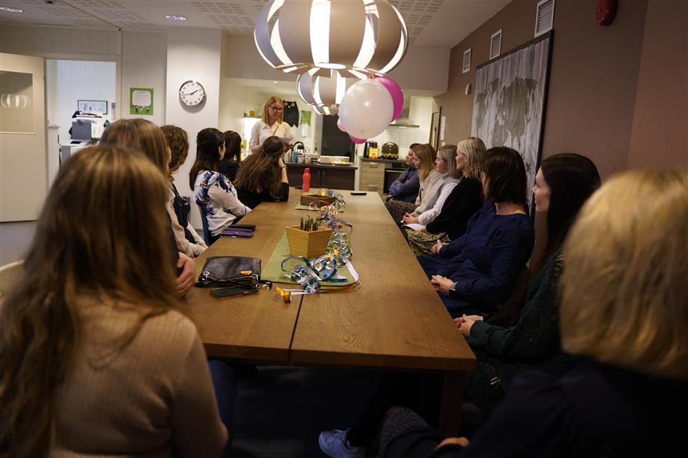 Mange mennesker som sitter rundt bordet inne på Ungdomskontakten, Sissel står ved enden og taler. - Klikk for stort bilde