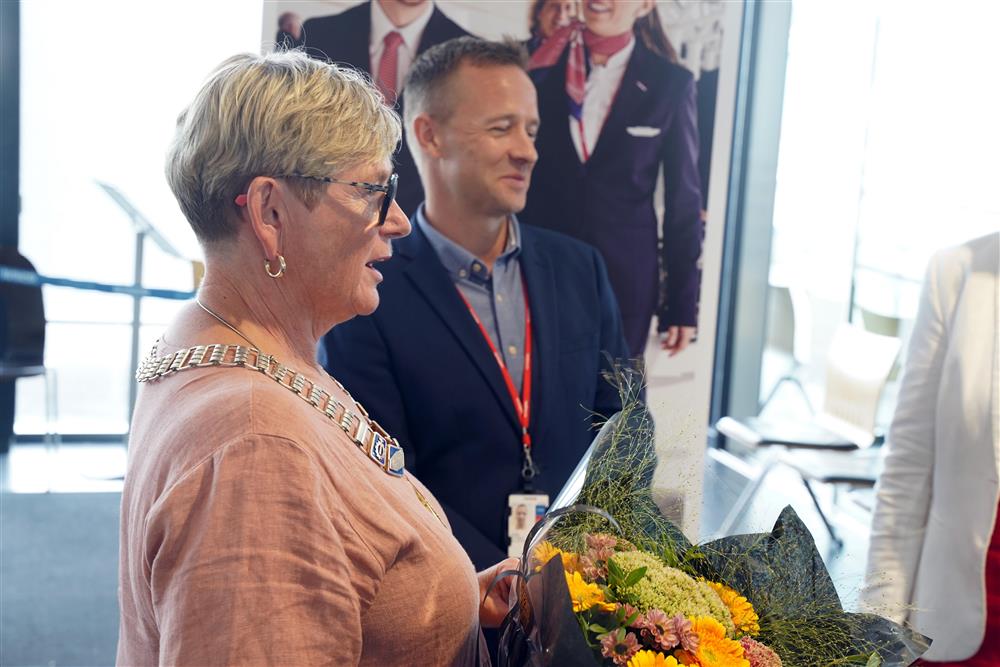 Ordfører Eli Arnstad som står med en blomsterbukett, i bakgrunnen en mann i dress. De er inne på flyplassen på Værnes. - Klikk for stort bilde