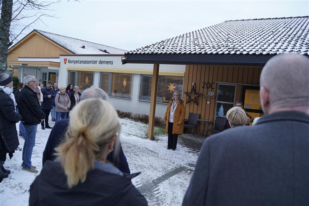 Flere mennesker som står utendørs og hører på talen til Venche Skjervold. Bak henne på veggen er skiltet til Kompetansesenter demens - Klikk for stort bilde