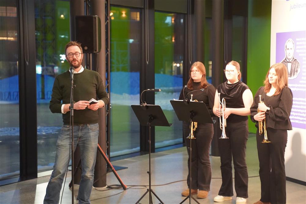 En mann som snakker i mikrofon foran tidslinjen til Ole Vig, rett bak ham står tre elever fra Kulturskolen klare til å spille instrumenter. - Klikk for stort bilde