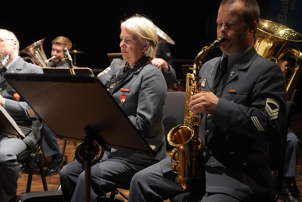 En mann og en dame i uniform som spiller instrumenter. Flere folk som spiller i bakgrunnen. - Klikk for stort bilde