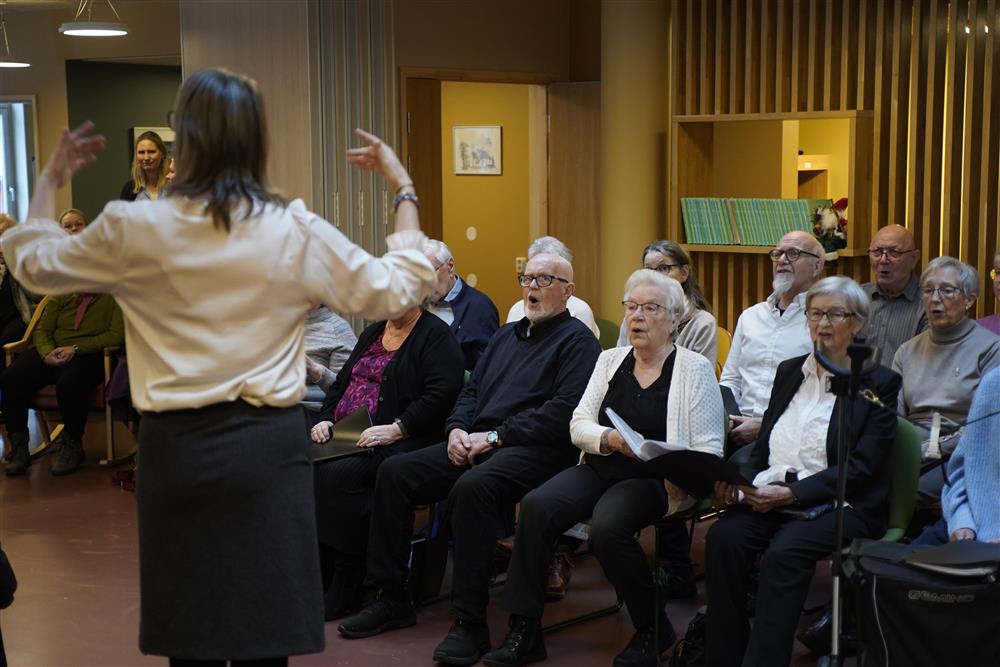 En dirigent som står foran mange eldre som deltar i demenskoret. De sitter i stoler inne i storstua til Fosslia omsorgsenter. - Klikk for stort bilde