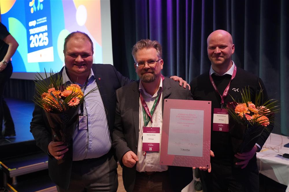 Tre menn som står sammen, en holder en pris og de to andre holder hver sin blomst. De står alle i dress. - Klikk for stort bilde