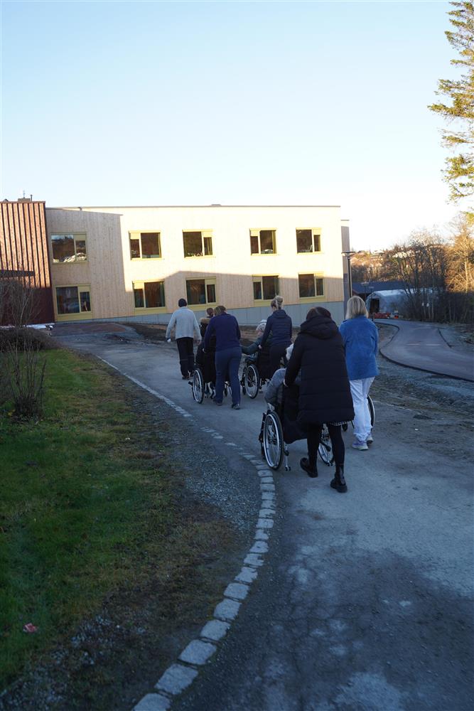 En lang rekke med pleiere som triller beboere i rullestoller til nye Fosslia Omsorgsenter - Klikk for stort bilde