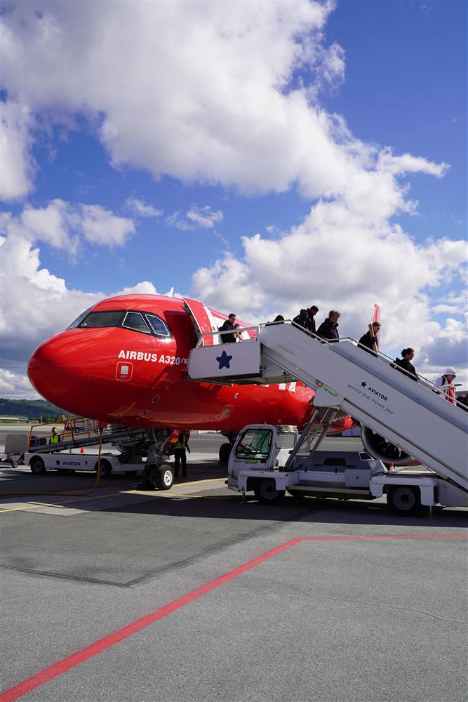 Et rødt fly i strålende sol, flere passasjerer sett fra siden på vei ned trappen fra flyet. - Klikk for stort bilde