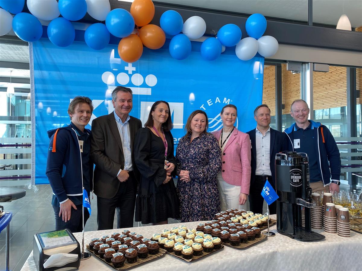 Syv mennesker foran KLM sin logovegg pyntet med ballonger og kake i forgrunnen - Klikk for stort bilde