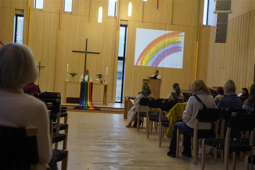 Regnbuemesse i Stjørdal kirke, en står på taler stolen, en prest står bak et bord med regnbue på. På veggen er det kors og en prosjektor viser bilde av en regnbue.  - Klikk for stort bilde