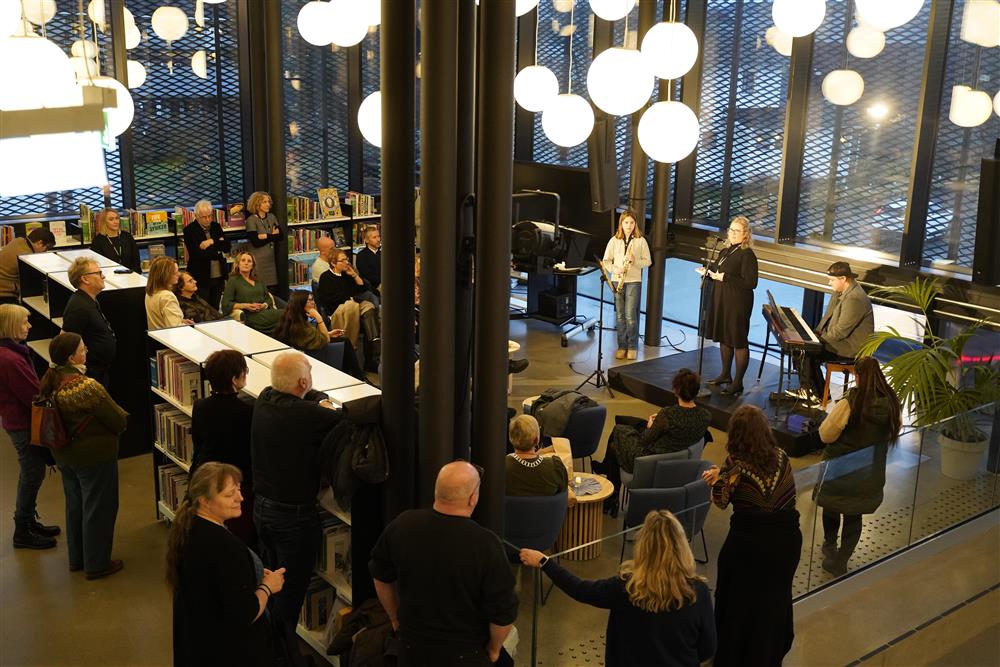 Mange mennesker som står rundt om i salongen, de følger med på biblioteksjefen som holder sin tale på scenen. To fra kulturskolen gjør seg klare bak på scenen. - Klikk for stort bilde