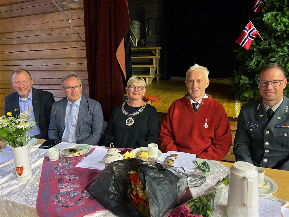 Lang bord med 6 personer. I midten ordføreren og Olaus Dalsplass. De er innendørs, bordet er dekket med kaffe og kaker. I bakgrunnen norske flagg og annen pynt. - Klikk for stort bilde